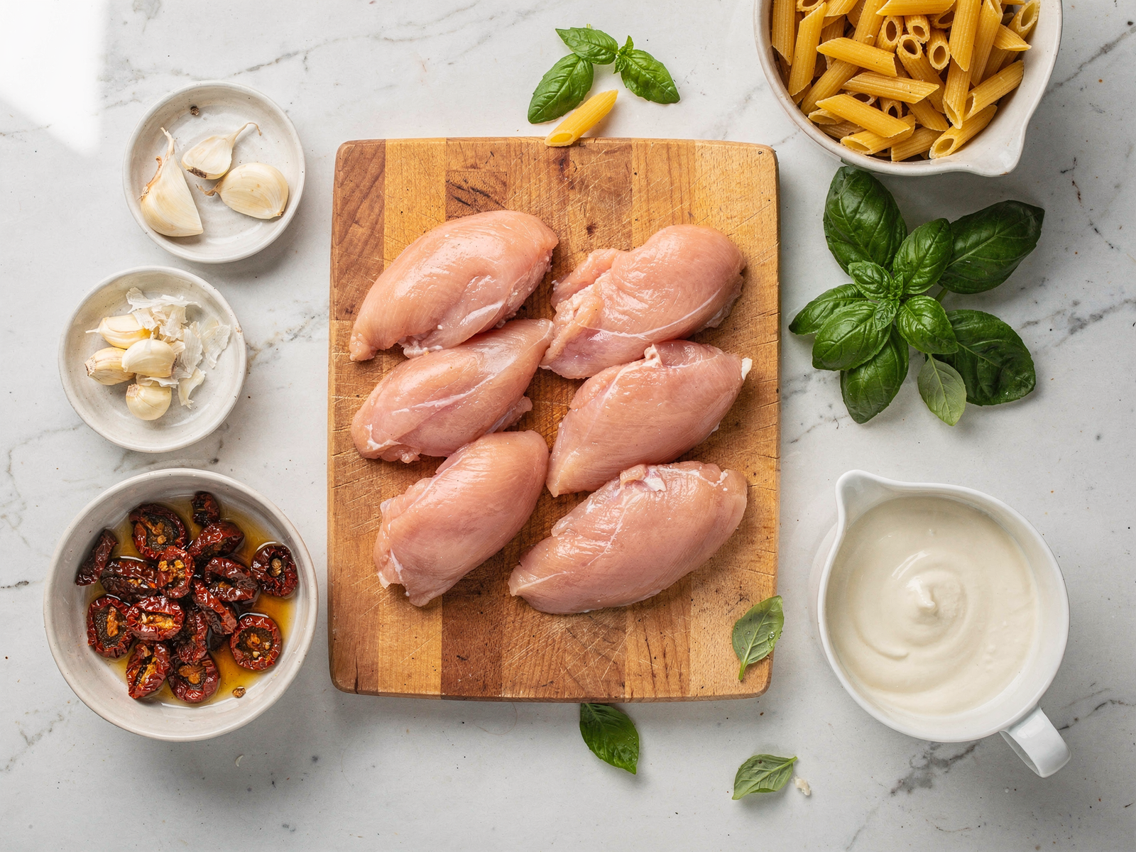 A marble countertop showing prepped ingredients for Marry Me chicken pasta one pot recipe including chicken breasts, garlic, sun-dried tomatoes, fresh basil, and cream