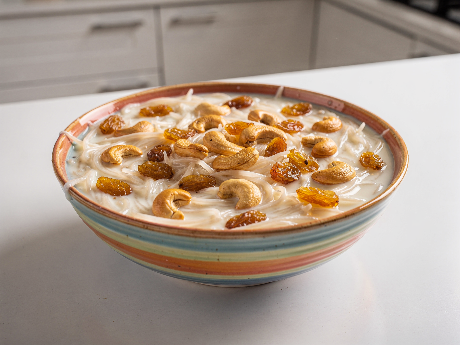 A colorful bowl of finished easy vermicelli milk dessert garnished with cashews and golden raisins on a white kitchen counter