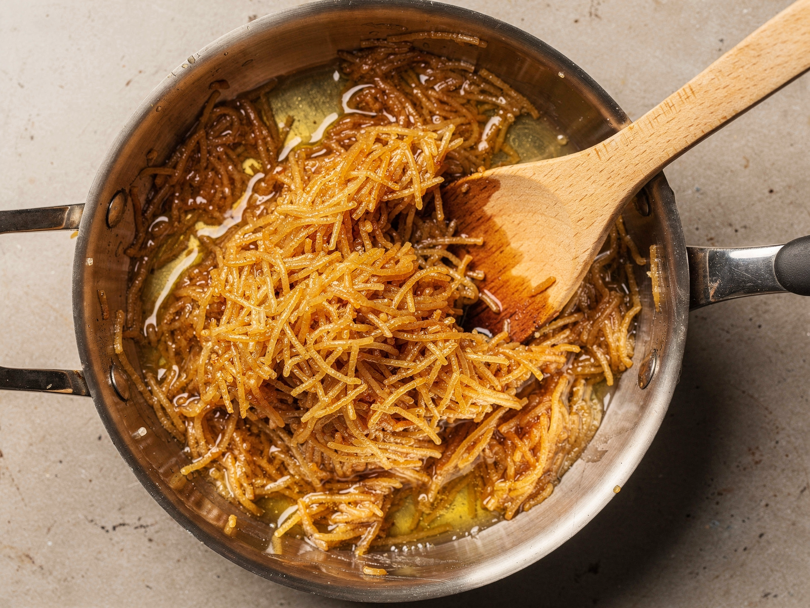 Golden-brown toasted vermicelli pasta pieces in ghee inside a stainless steel saucepan with a wooden spoon